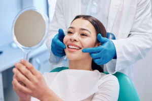 Smiling woman sitting in a dental chair holding a mirror, while a dentist wearing blue gloves gently checks her teeth.